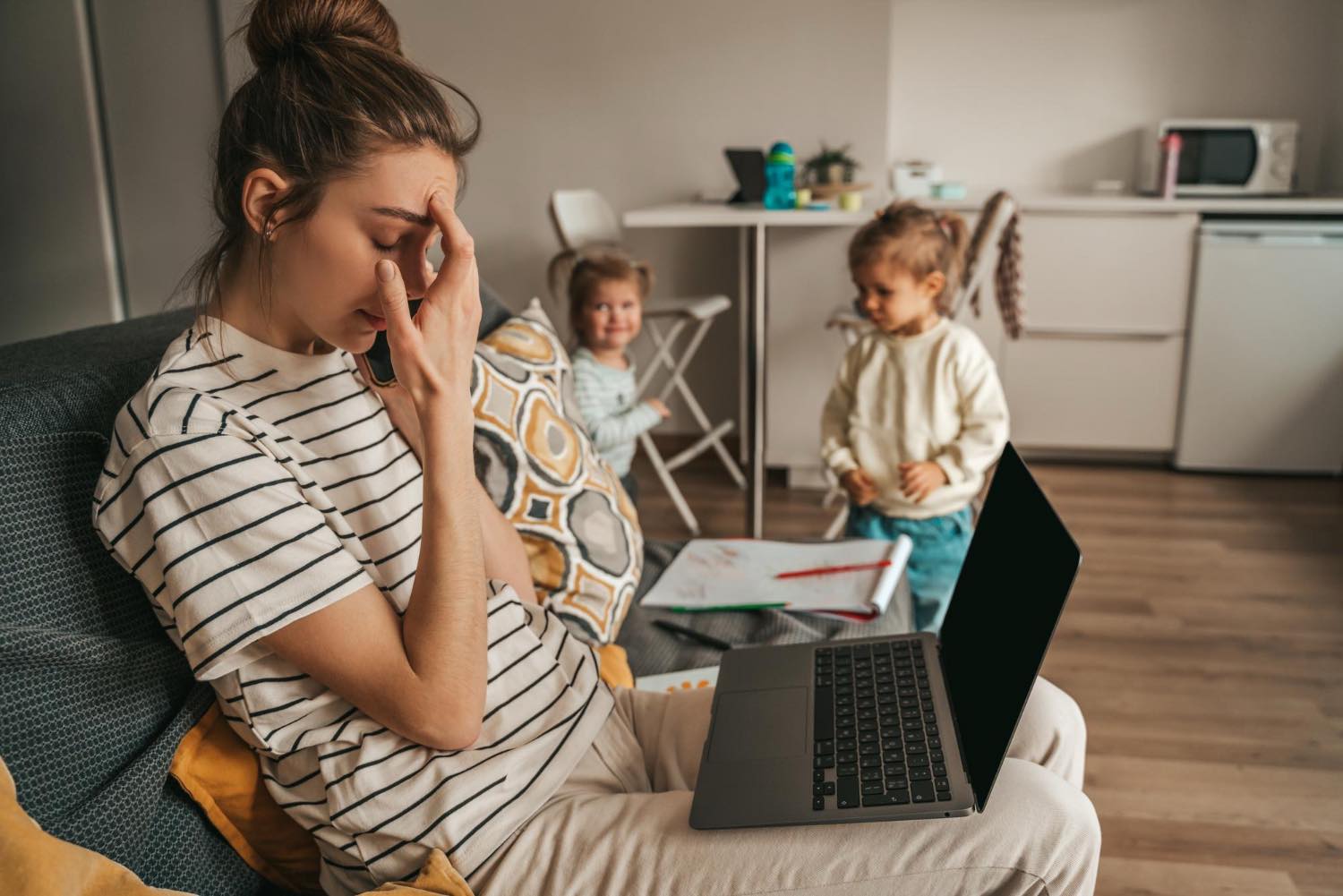 Stressed mother with hand on head sitting near two young children, illustrating the mental load of parenting and anxiety