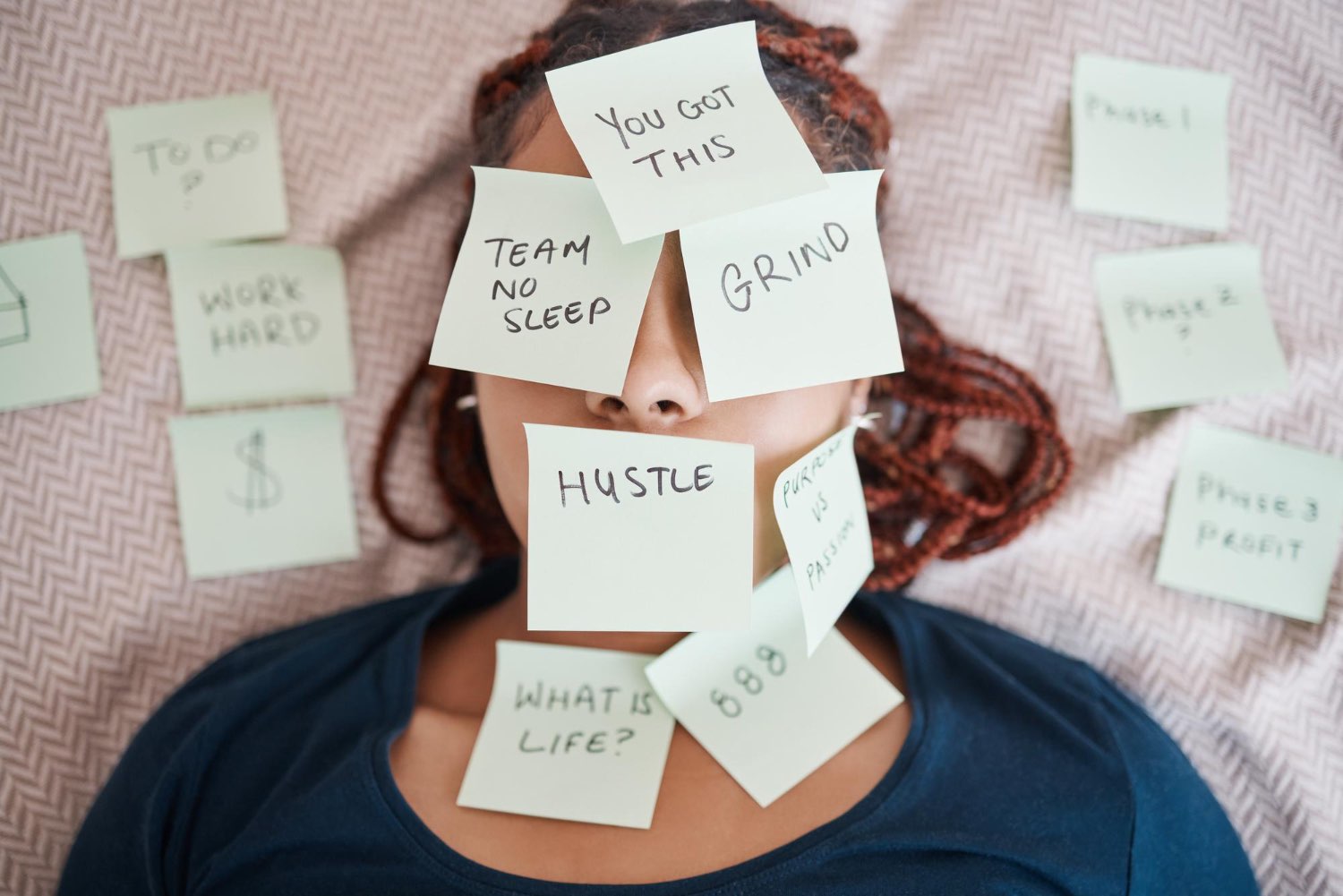 Woman lying down with sticky notes covering her face reading "hustle", "grind" and "work hard", representing the mental overwhelm of anxiety and stress