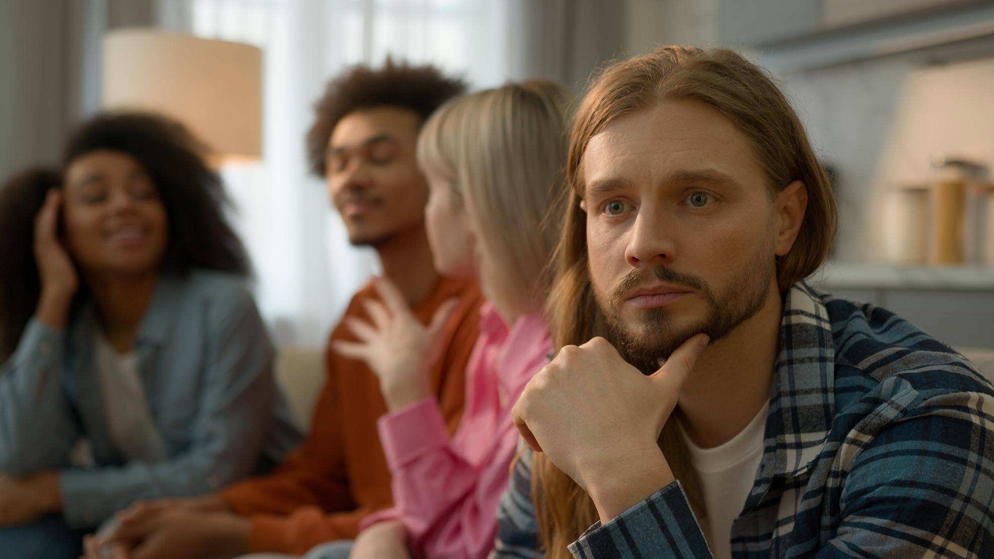 Chronic anxiety and worry, man with hand on chin looking distant and preoccupied while friends socialise in the background, illustrating social withdrawal driven by persistent worry